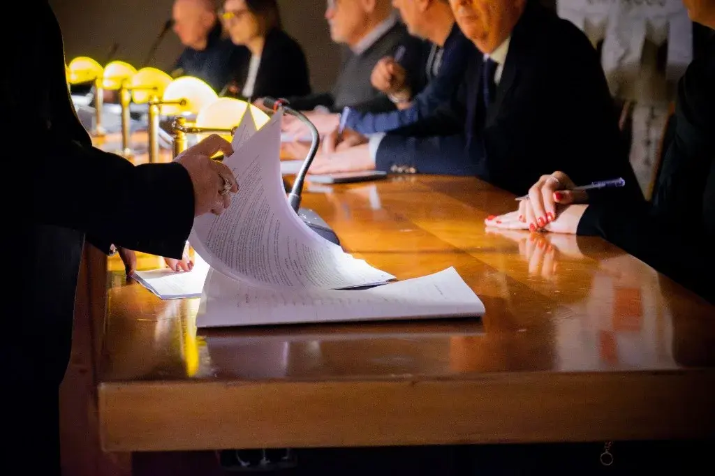 Hands reviewing formal documents at a hearing table with a panel seated opposite